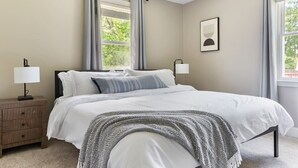Et soveværelse med en seng, to sengeborde, en lampe, et indrammet billede og et vindue med gardiner.