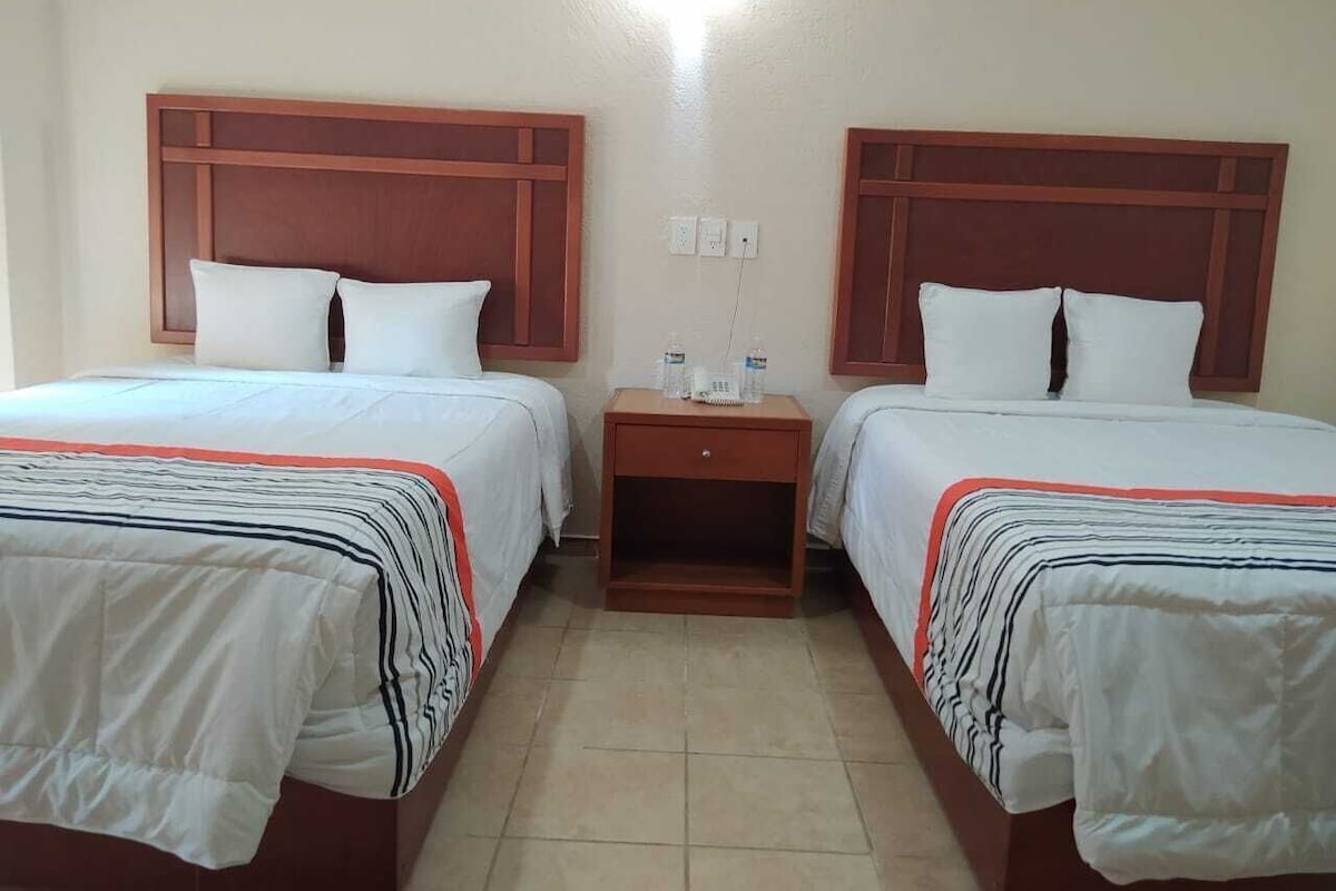 A hotel room with two beds, each with a wooden headboard, a small wooden nightstand between them, and a light fixture above.