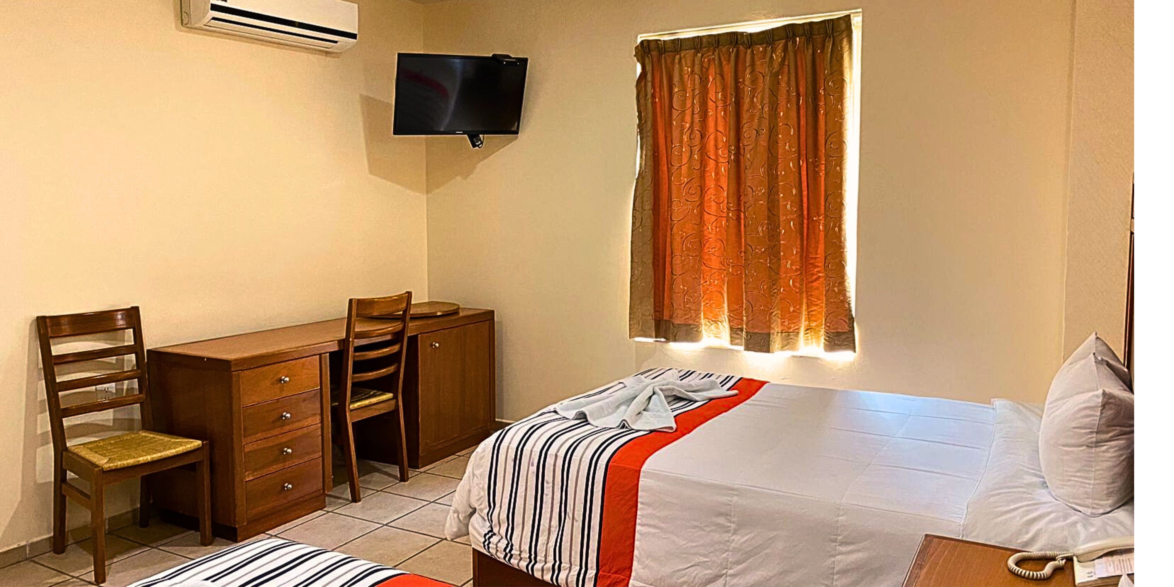 A hotel room with a bed, a desk, a chair, a TV mounted on the wall, and a window with curtains.