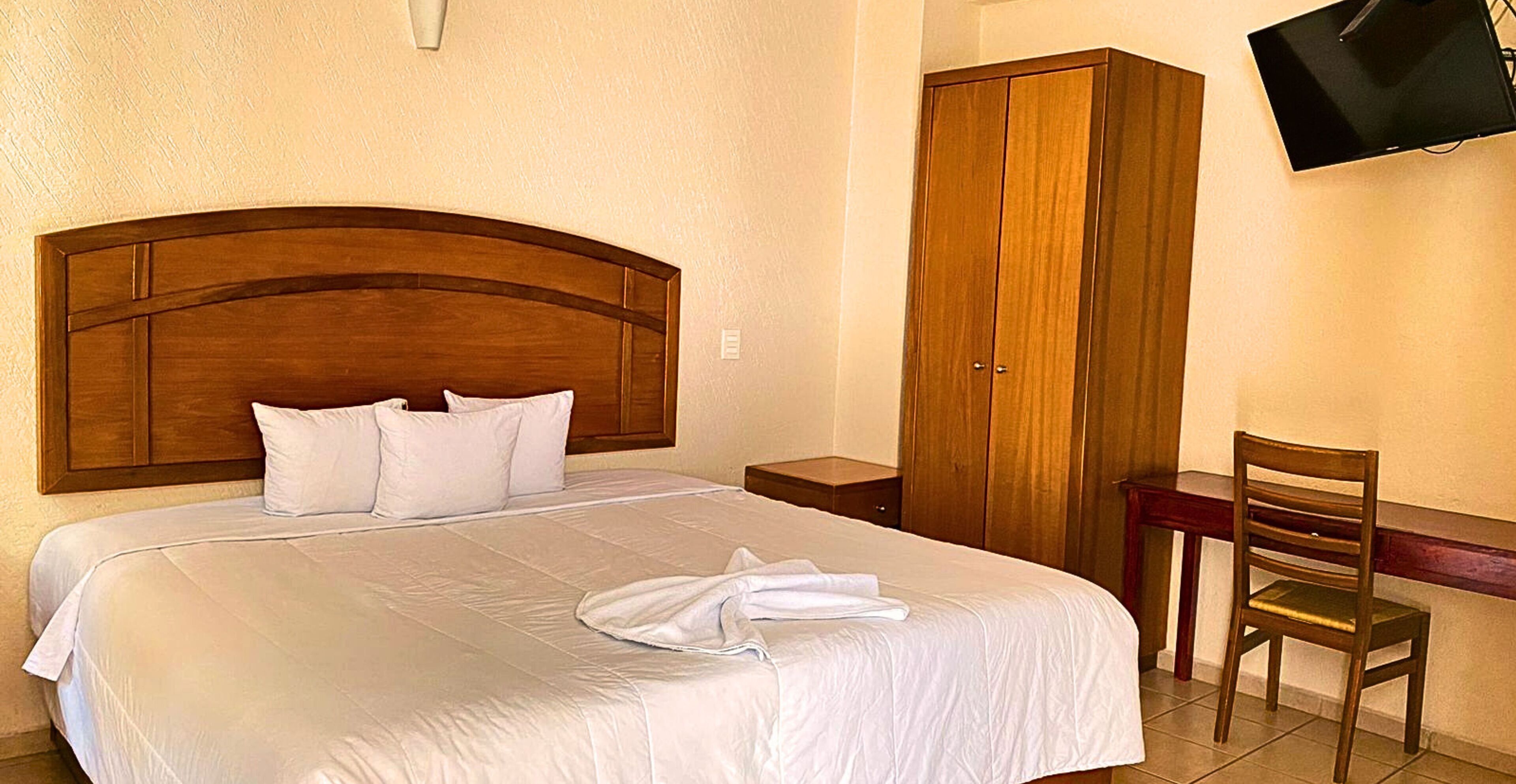 Uma cama bem arrumada com lençóis brancos, cabeceira de madeira, guarda-roupa de madeira, televisão na parede e uma cadeira de madeira.