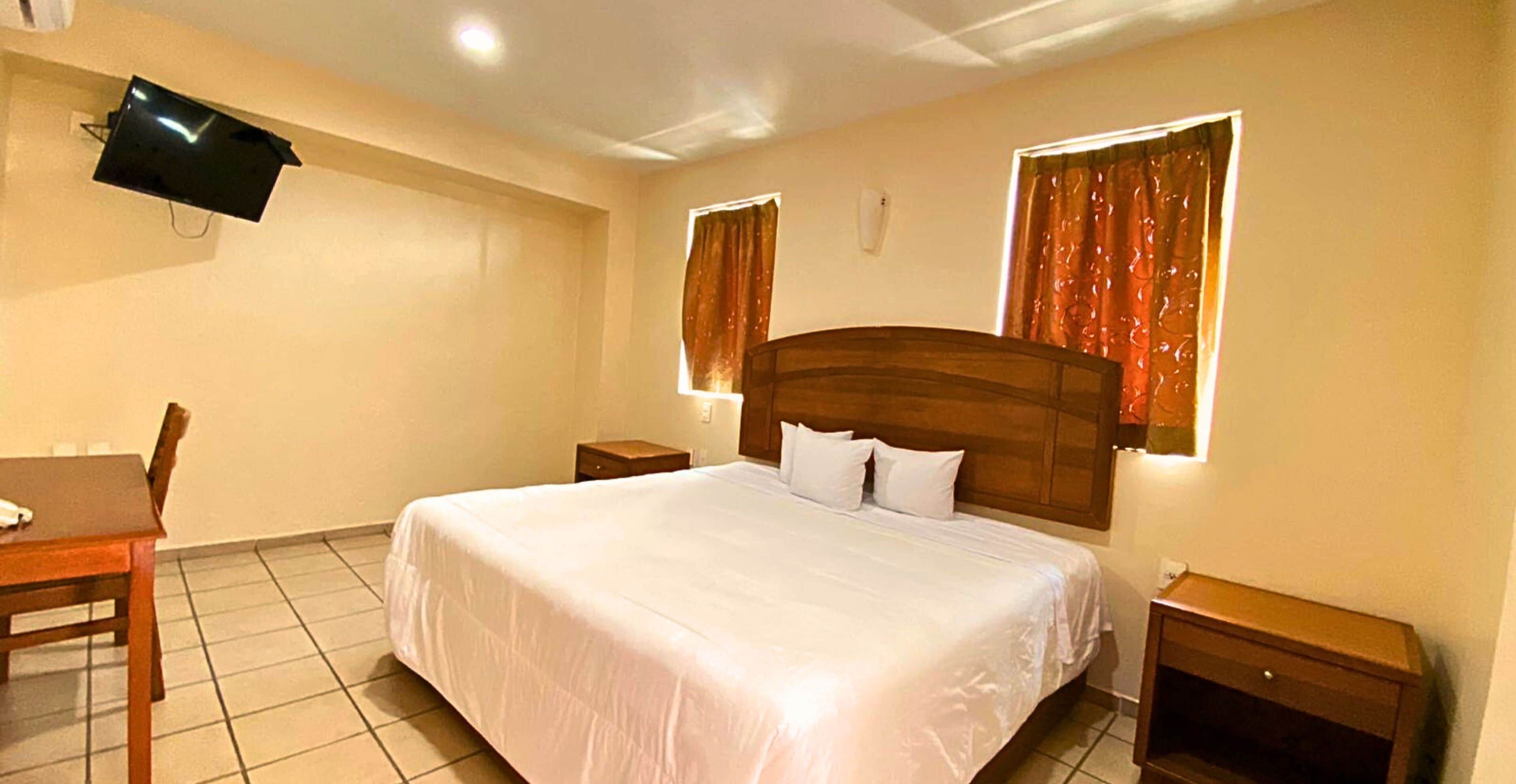 Quarto de hotel com uma cama grande, uma escrivaninha e uma televisão fixada na parede.
