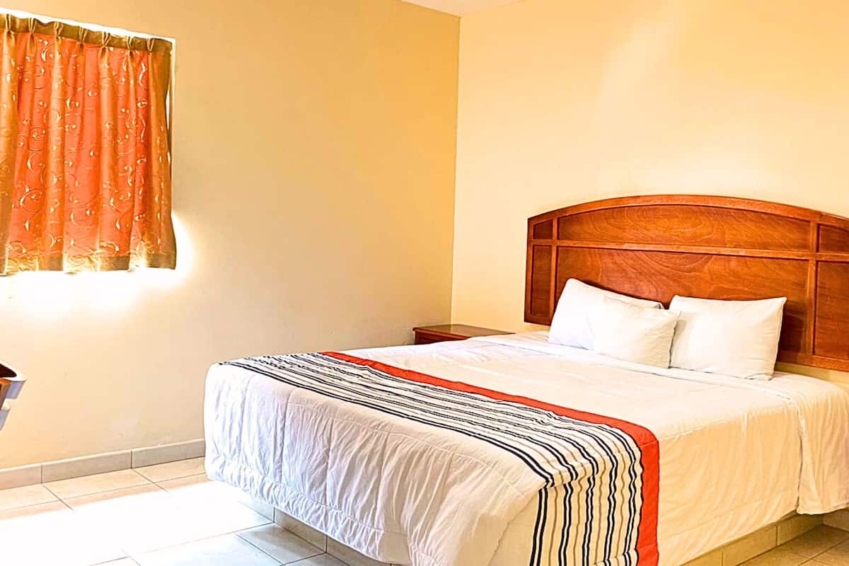A hotel room with a large bed, wooden headboard, white bedding, a bedside table with a lamp, and a window with curtains.