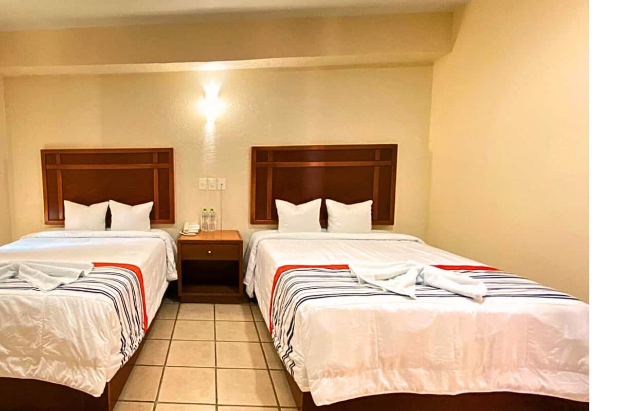 A hotel room with two beds, each with a wooden headboard, white bedding, and striped blankets. There is a nightstand with a lamp and a phone between the beds.