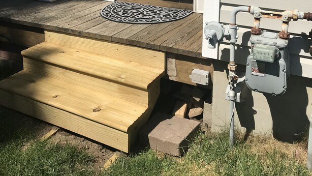 Eine Holzterrasse mit einer Bank, einem kleinen Teppich und einem Technikschrank neben einem Haus mit weißer Fassade und einer Tür.