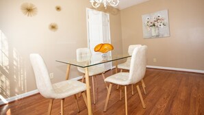 Un comedor con una mesa de vidrio, cuatro sillas blancas, una lámpara de araña y un cuadro en la pared.