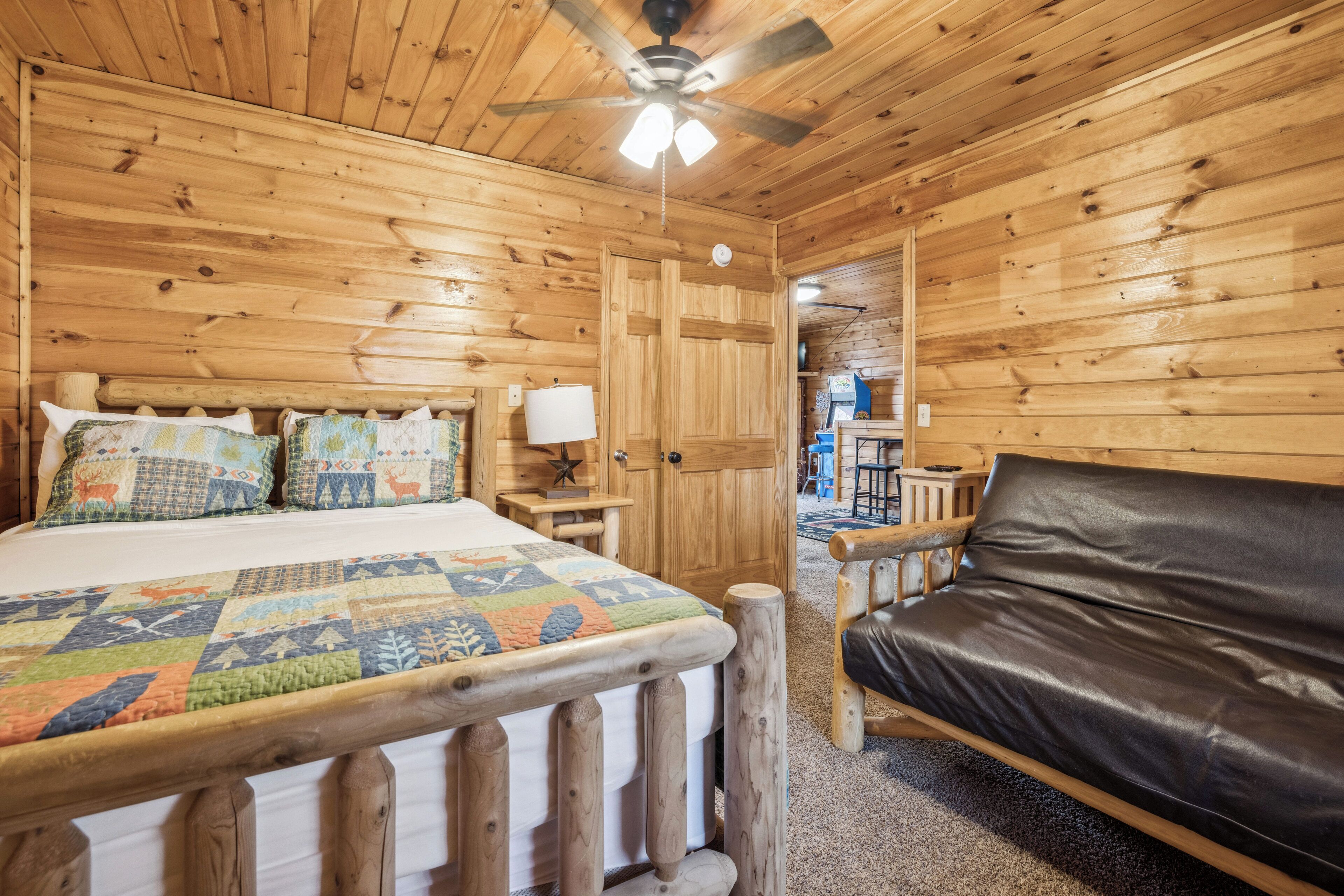 Un dormitorio de cabaña de madera con cama, sofá de cuero negro, ventilador de techo y puerta de madera que da a una cocina.