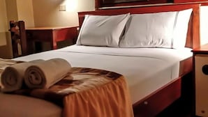 Una cama bien hecha con sábanas blancas y dos almohadas, una mesita de noche de madera con una lámpara, y una toalla enrollada en el borde de la cama.