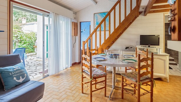 A cozy dining area with wooden chairs and a table set for four, a staircase leading to an upper level, and a television on a stand. - Maison aux Oiseaux - Welkeys (Dolus-d'Oléron)