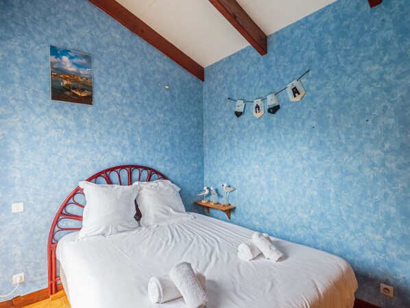 A bedroom with a bed, white bedding, a red metal headboard, a wall-mounted picture, and a small shelf with towels and a bird figurine. - Maison aux Oiseaux - Welkeys (Dolus-d'Oléron)