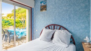 A bedroom with a bed, a wooden nightstand, and a view through a glass door to an outdoor area with a table and chairs. - Maison aux Oiseaux - Welkeys (Dolus-d'Oléron)
