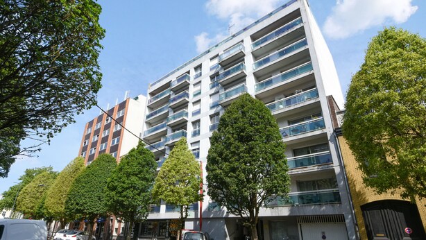 Un immeuble résidentiel à plusieurs étages, doté de balcons, avec des voitures garées et des arbres le long de la rue.