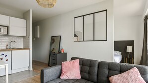 Un salon moderne avec un canapé gris, deux coussins roses, un luminaire suspendu en fibres tressées, un grand miroir et un coin cuisine doté de meubles blancs et d’un réfrigérateur blanc.