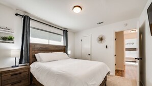 Un dormitorio con cabecera de madera, ropa de cama blanca, una mesita de noche con lámpara, una ventana con cortinas y una puerta que da a otra habitación.