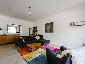 A modern living room with a black sofa, a wooden coffee table, a colorful rug, and a white fluffy dog. - Appartement Mésangia - Welkeys (Villeneuve-Loubet)