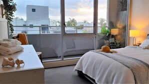 Een hotelkamer met een groot raam, een bed met witte lakens, een bureau met handdoeken en een plant op het nachtkastje.