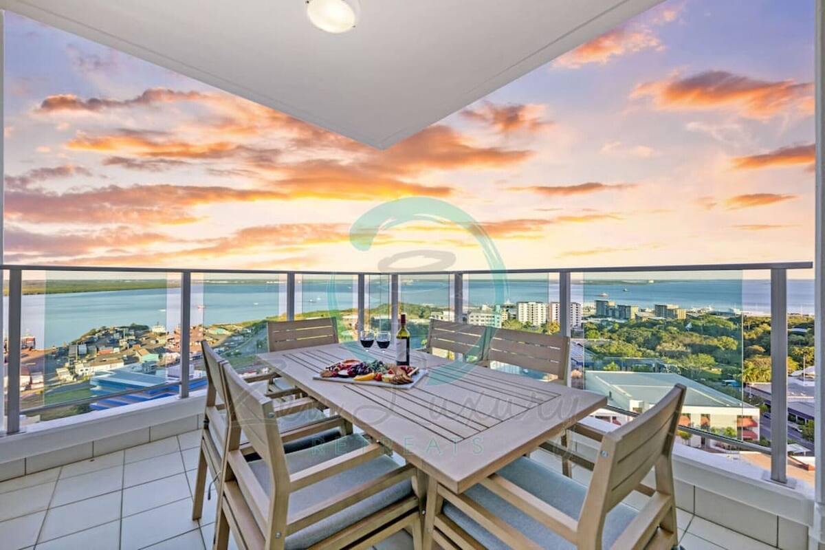 A balcony with a table set for dining, overlooking a coastal cityscape with a sunset view.