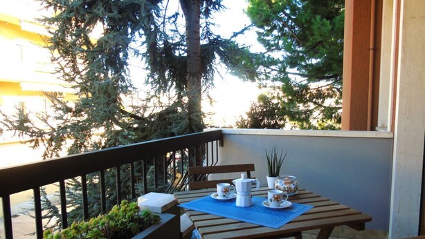 A balcony with a wooden table set for two, a potted plant, and a view of trees and a building. - Medusa - Ground floor apartment, a few steps from the sea and the center (Pineto)