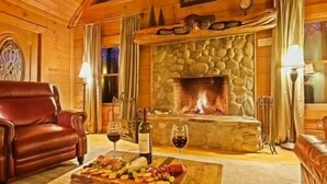 Interior acogedor de una cabaña de troncos con chimenea de piedra, un sillón de cuero y una bandeja de frutas sobre una mesa de madera.