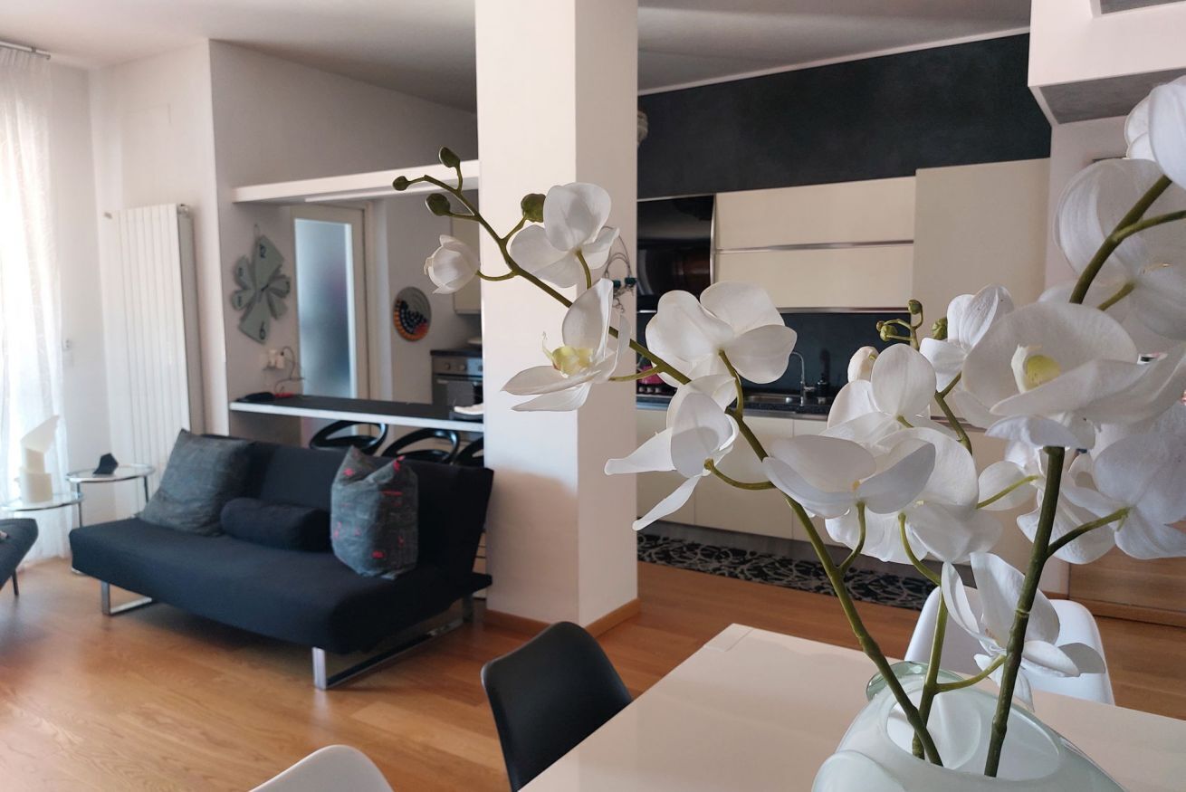 Ein modernes Wohnzimmer mit einer schwarzen Couch, einem Esstisch und weißen Orchideen in einer Vase.
