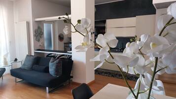 Ein modernes Wohnzimmer mit einer schwarzen Couch, einem Esstisch und weißen Orchideen in einer Vase.