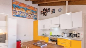 A compact kitchen with white and yellow cabinets, a wooden dining table, and a painting above the sink.