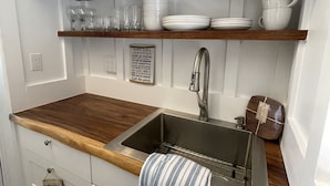 Una cocina con encimera de madera, pileta de acero inoxidable y muebles blancos. Hay estantes sobre la pileta con platos y vasos.