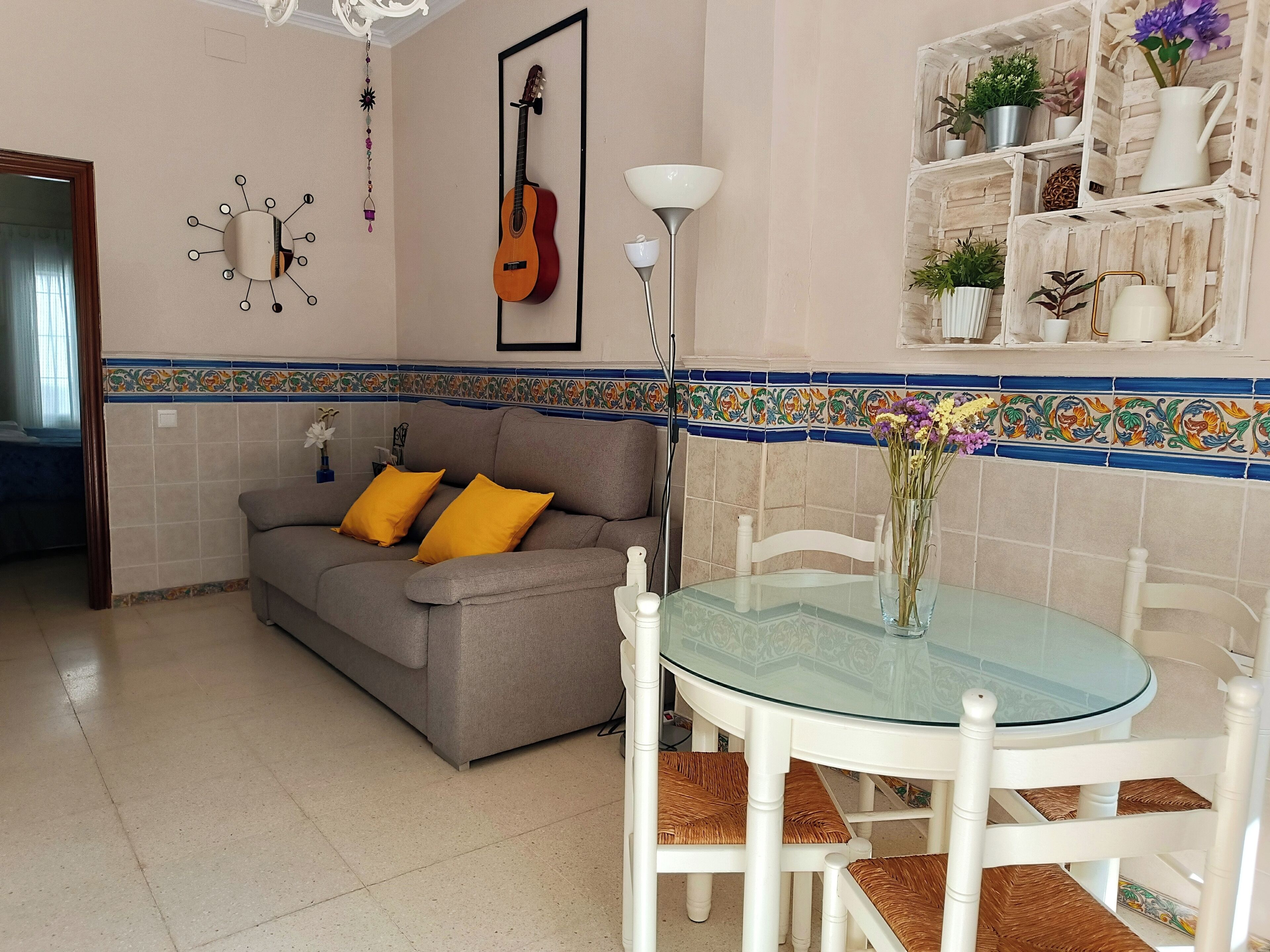 A living room with a grey sofa, yellow cushions, a glass-top dining table, white chairs, a guitar on the wall, and a decorative shelf with plants.