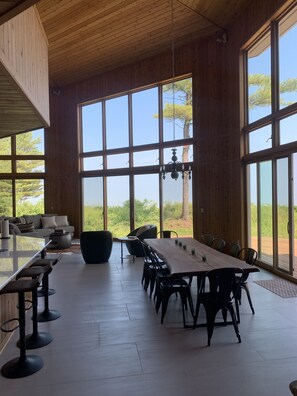 Un vaste espace dédié à la salle à manger, avec une longue table et des chaises, de grandes fenêtres offrant une vue sur l’extérieur et un lustre suspendu.