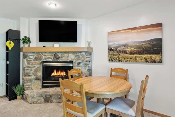 A living room with a stone fireplace, a round wooden dining table, and a flat-screen TV mounted above the fireplace.