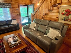 A wooden cabin interior with a sofa, a coffee table with remote controls, and a staircase.