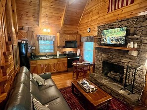 A cozy cabin interior with a stone fireplace, wooden walls, a leather sofa, a dining area, and a kitchen with a refrigerator and stove.