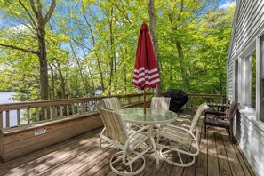 A wooden deck with a round table and chairs, a red-striped umbrella, and a view of a lake surrounded by trees. - Pond Retreat: Beach, Kayak, Canoe, Deck, Firepit, (New Durham)
