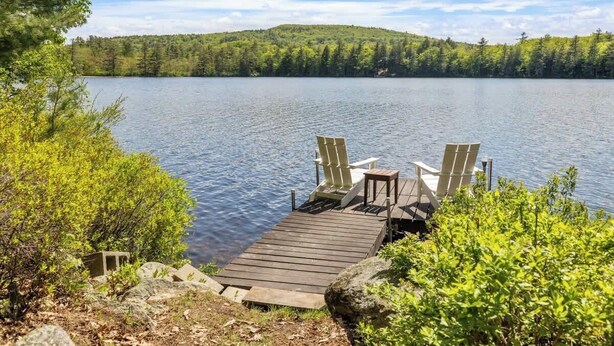 Une jetée en bois, agrémentée de deux fauteuils Adirondack et d’une petite table, offre une vue sur un lac paisible, avec une colline boisée en arrière-plan.