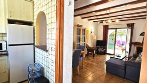 Eine Küche mit Kühlschrank, Mikrowelle und einem kleinen Tisch mit einem Korb. Es gibt einen Essbereich mit Tisch und Stühlen sowie ein Wohnzimmer mit Sofa und Fernseher.
