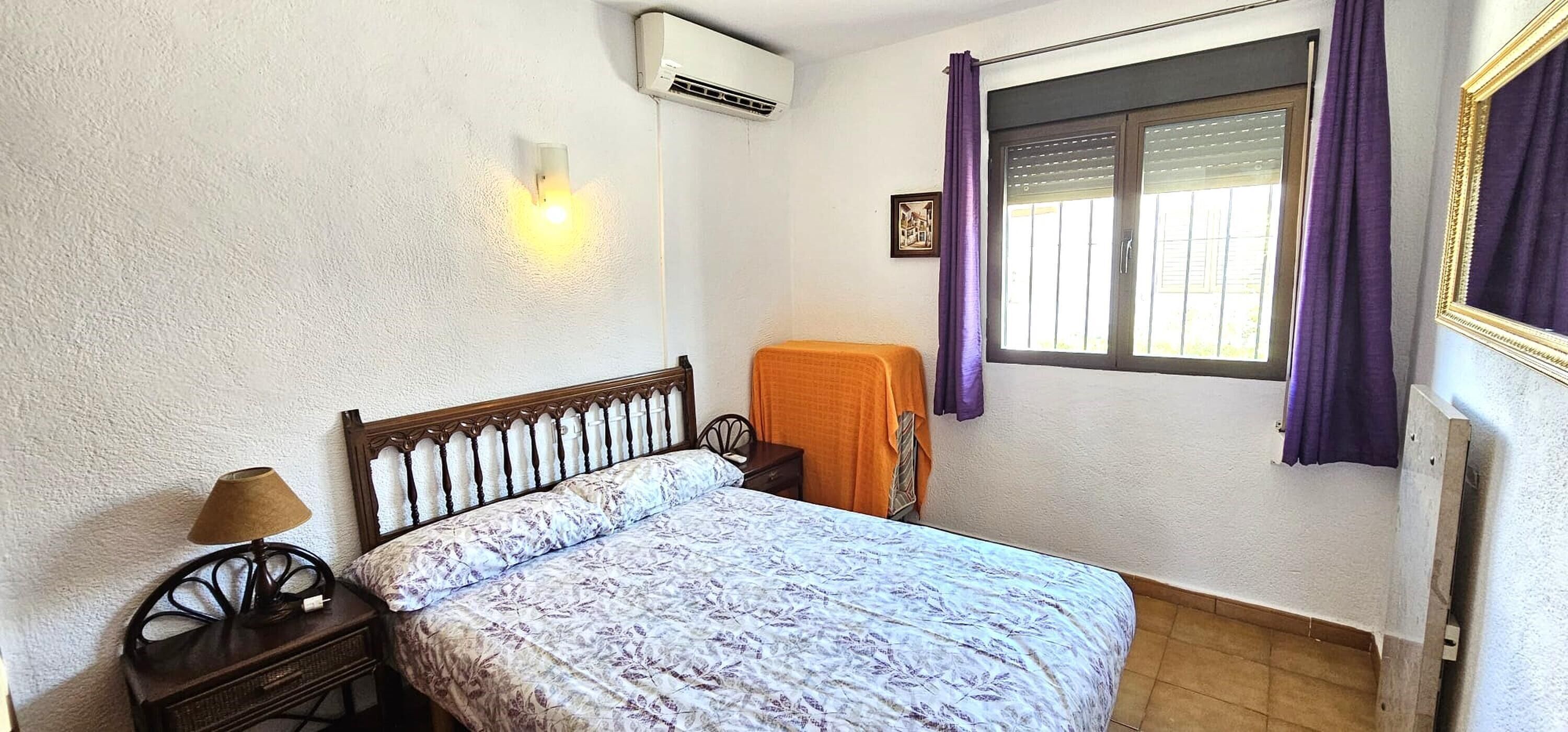 Un dormitorio con cama, mesita de noche, lámpara, ventana con cortinas y un equipo de aire acondicionado.