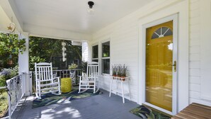 Een veranda met witte relingen, twee schommelstoelen, een gele lantaarn en een deur met een geel glasraam.