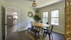 Een eetruimte met een ronde tafel, zwarte stoelen, een geweven bank, een hanglamp en een raam met uitzicht op bomen.