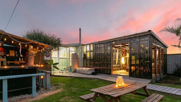 Un entorno rústico al aire libre con una mesa de picnic de madera, un cobertizo de metal y una zona de bar con luces colgantes.