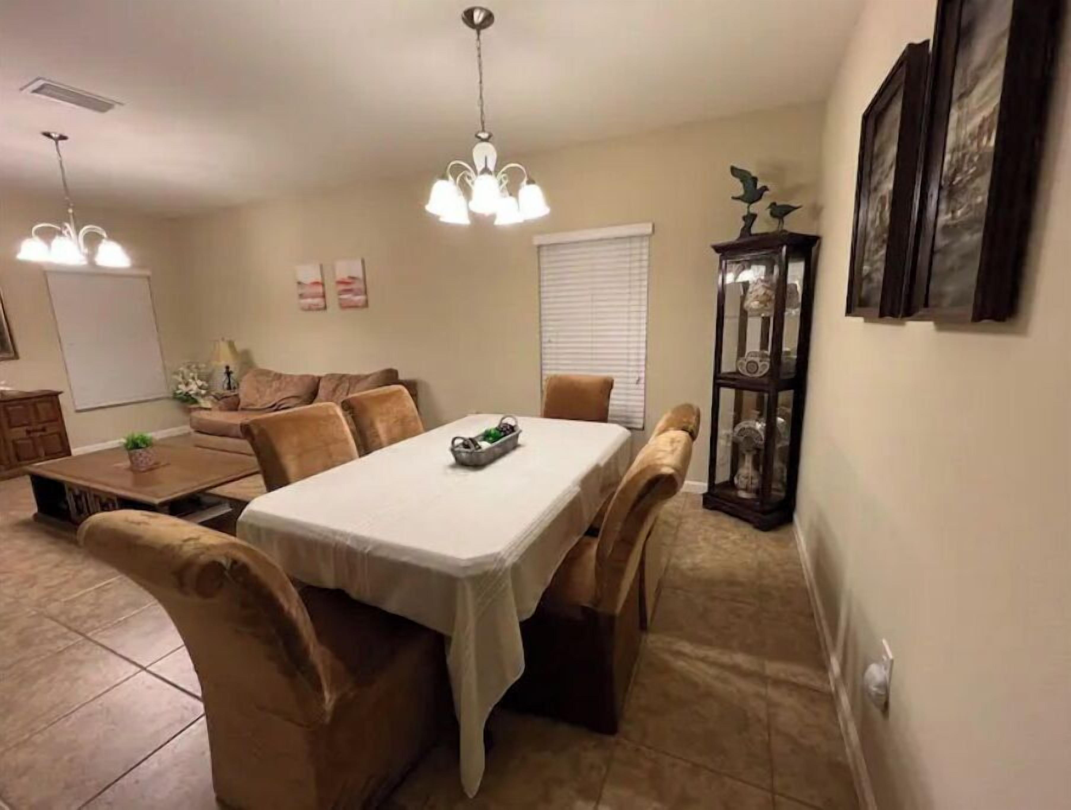 Un coin repas avec une table, des chaises et un lustre. On aperçoit un canapé et un meuble orné d’objets décoratifs à l’arrière-plan.