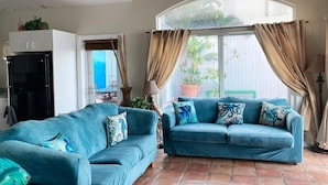 A living room with two blue sofas, a ceiling fan, a refrigerator, and a large window with curtains. - Ocean views from every room! (St Thomas)