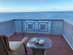 A balcony with a glass table and a wicker chair, overlooking the ocean.