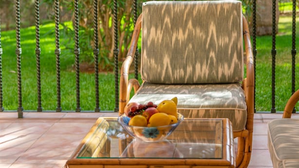 Une chaise en osier dotée d’un siège et d’un dossier rembourrés, placée sur une table en verre sur laquelle se trouve un saladier rempli de fruits.