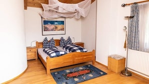 Une chambre avec un lit en bois, des coussins à carreaux, un tapis bleu et rouge, un tableau encadré au mur et une fenêtre avec des rideaux à motifs.