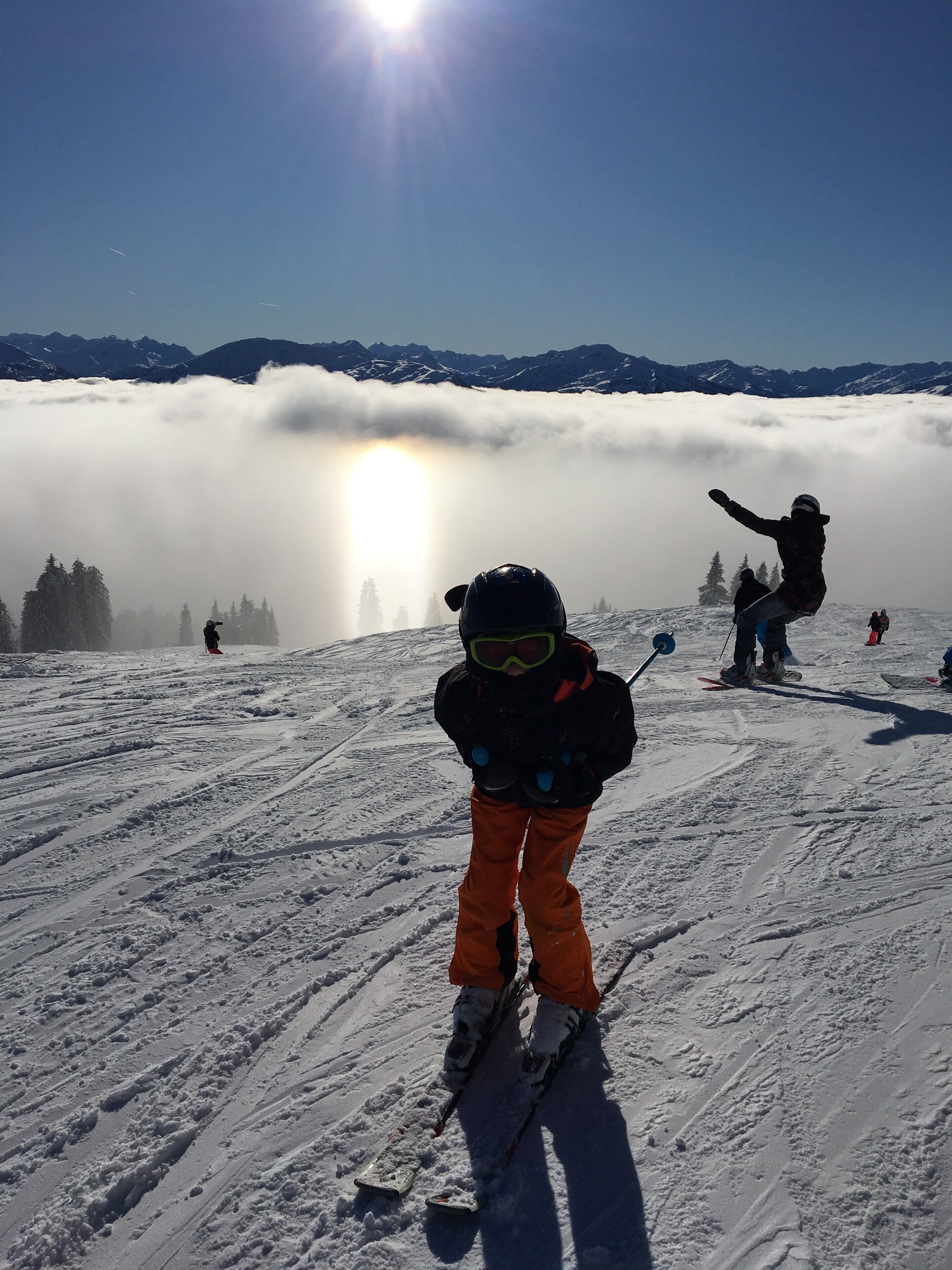 A snowy ski slope with skiers and a snowboarder, mountains in the background, and a sun shining through clouds.