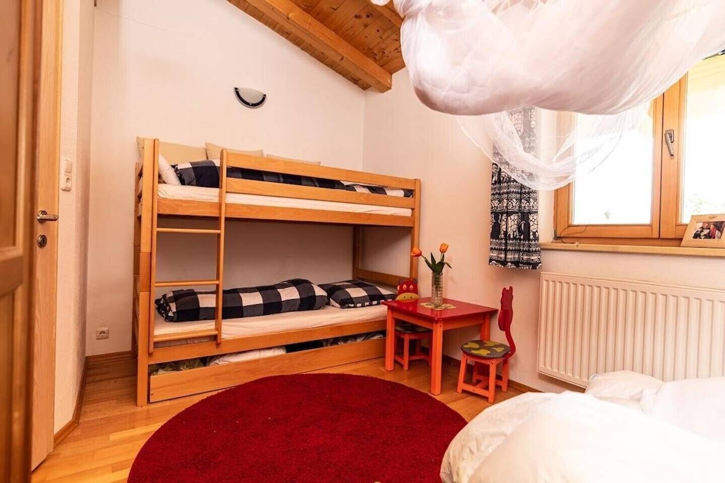 Ein Schlafraum mit Etagenbetten, Holzboden, einem roten Teppich, einem kleinen Tisch mit Stuhl und einem Fenster mit Vorhängen.