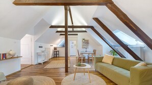 Habitación en el ático con un sofá verde, un tapete redondo, vigas de madera, una zona de comedor y una cocina al fondo.