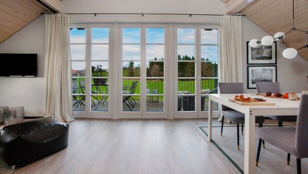 A spacious room with a large sliding glass door leading to an outdoor patio, a dining table with chairs, a black leather ottoman, and a mounted flat-screen TV. - Appartement with Balcony and Wi-Fi (Wangen im Allgäu)