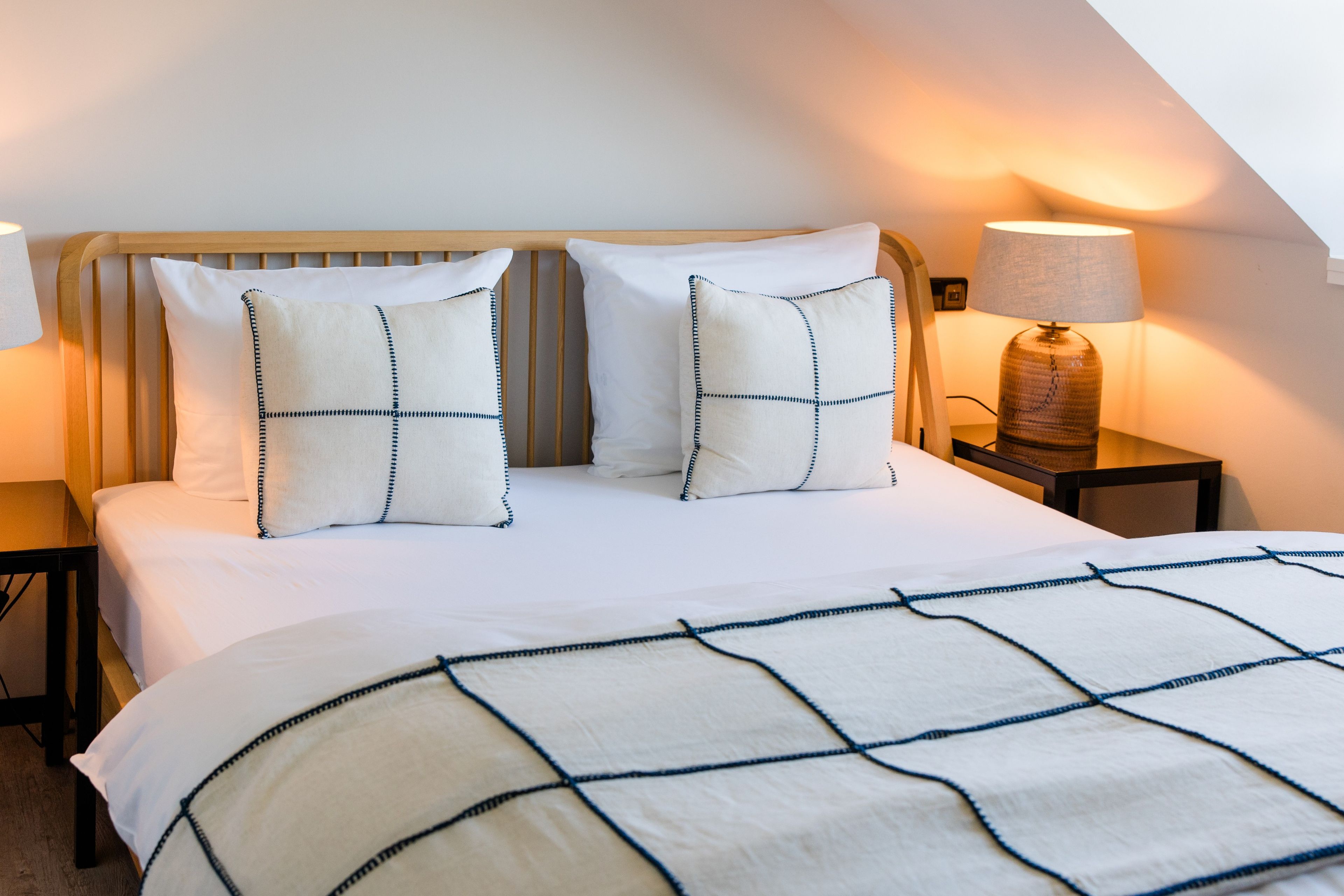 Una cama bien hecha con colcha blanca y azul a cuadros, dos almohadas con un diseño azul y blanco, y una lámpara de noche sobre una mesita de madera.