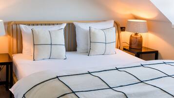 Una cama bien hecha con colcha blanca y azul a cuadros, dos almohadas con un diseño azul y blanco, y una lámpara de noche sobre una mesita de madera.
