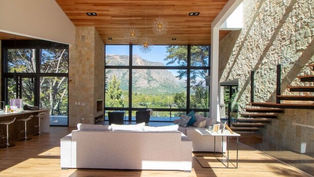 A spacious living area with a large white sofa, a dining bar, and a stone wall with a view of mountains. - New Modern Home, Golf, Polo, Few Minutes From Catedral Ski Resort.  (San Carlos de Bariloche)